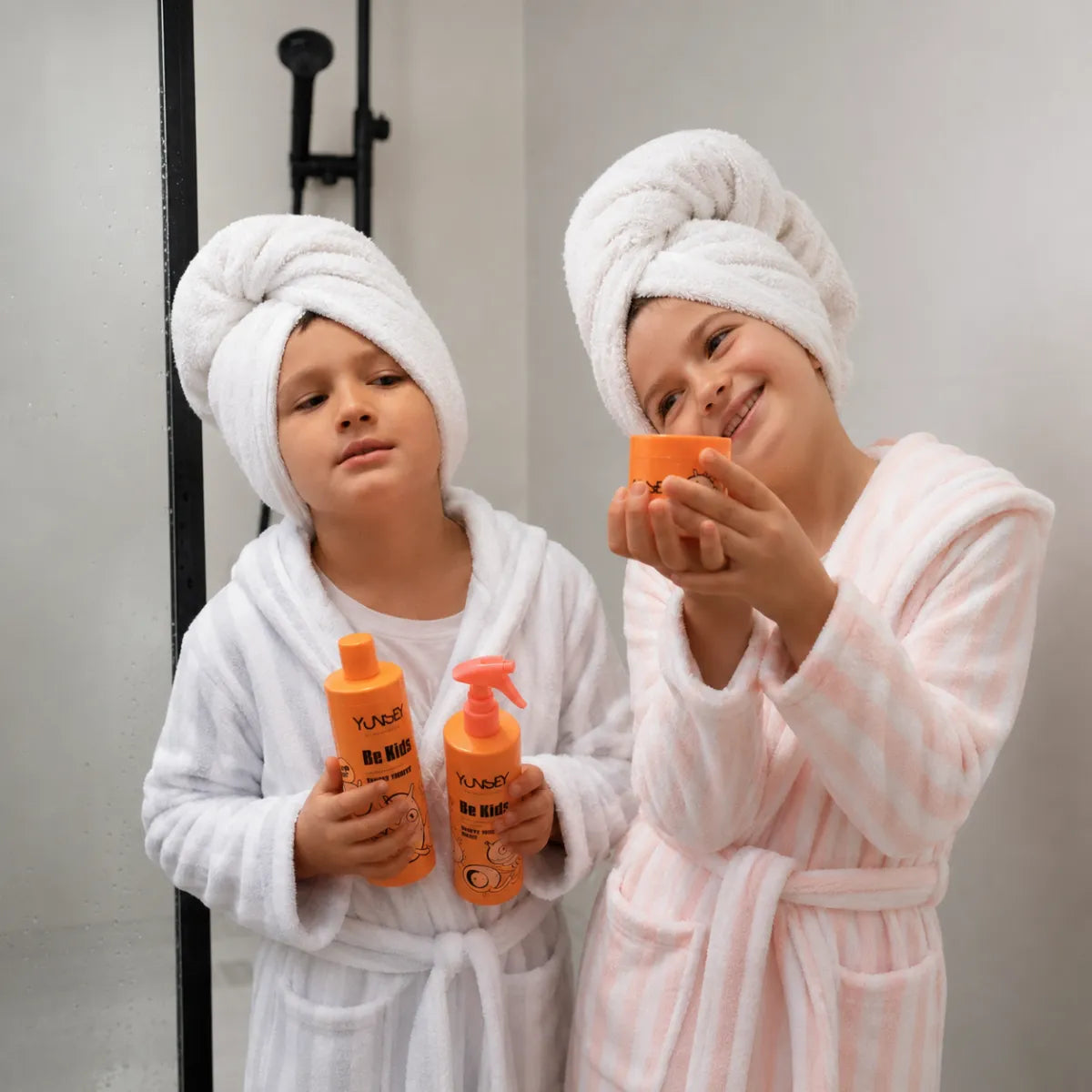 Dos niños con albornoces sostienen el champú, acondicionador y cera del pack Yunsey Be Kids para el cuidado del cabello infantil en el baño.